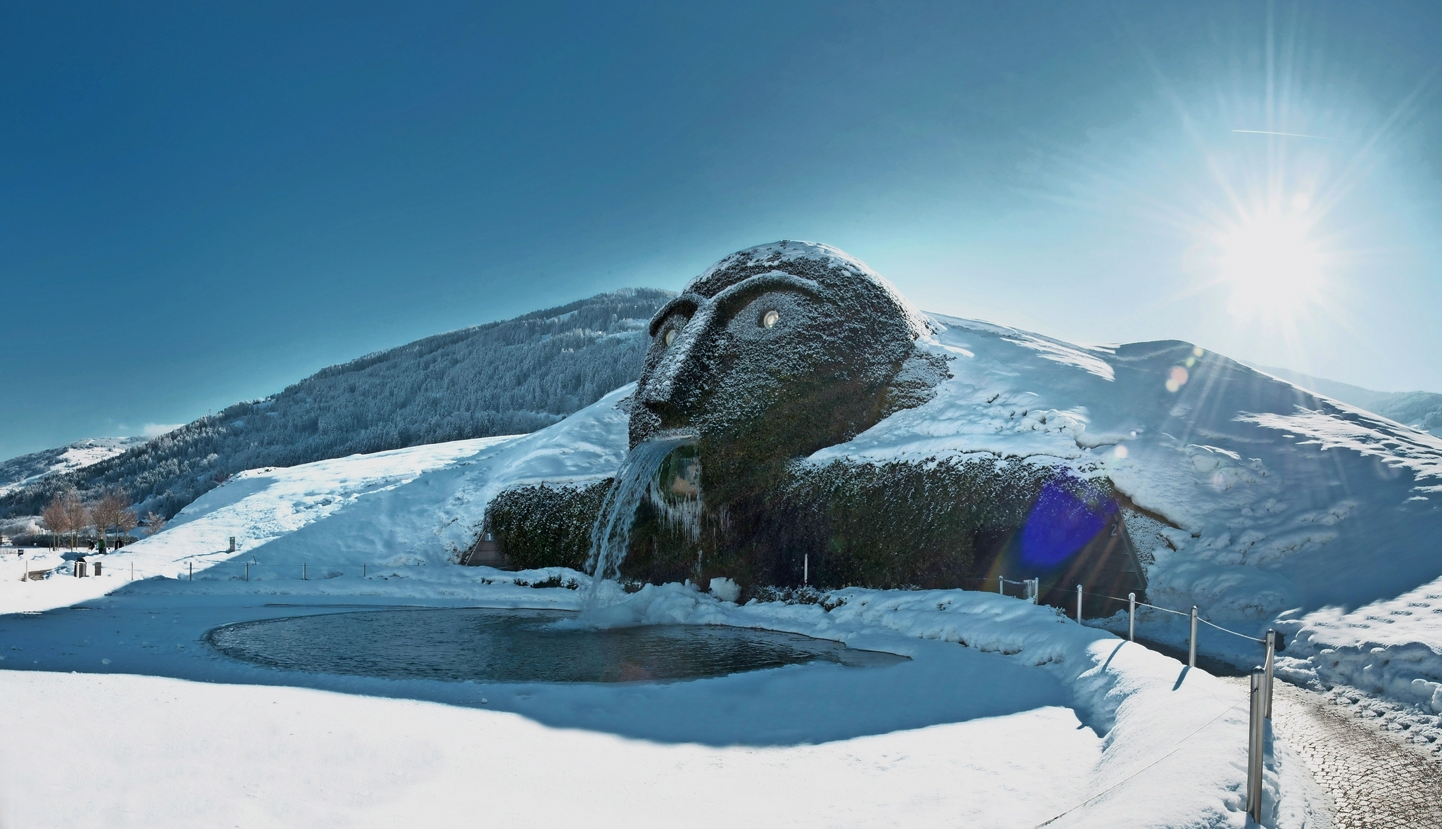 Schneebedeckter Riesenkopf mit Wasserspeier und Teich vor schneebedecktem Hügel.